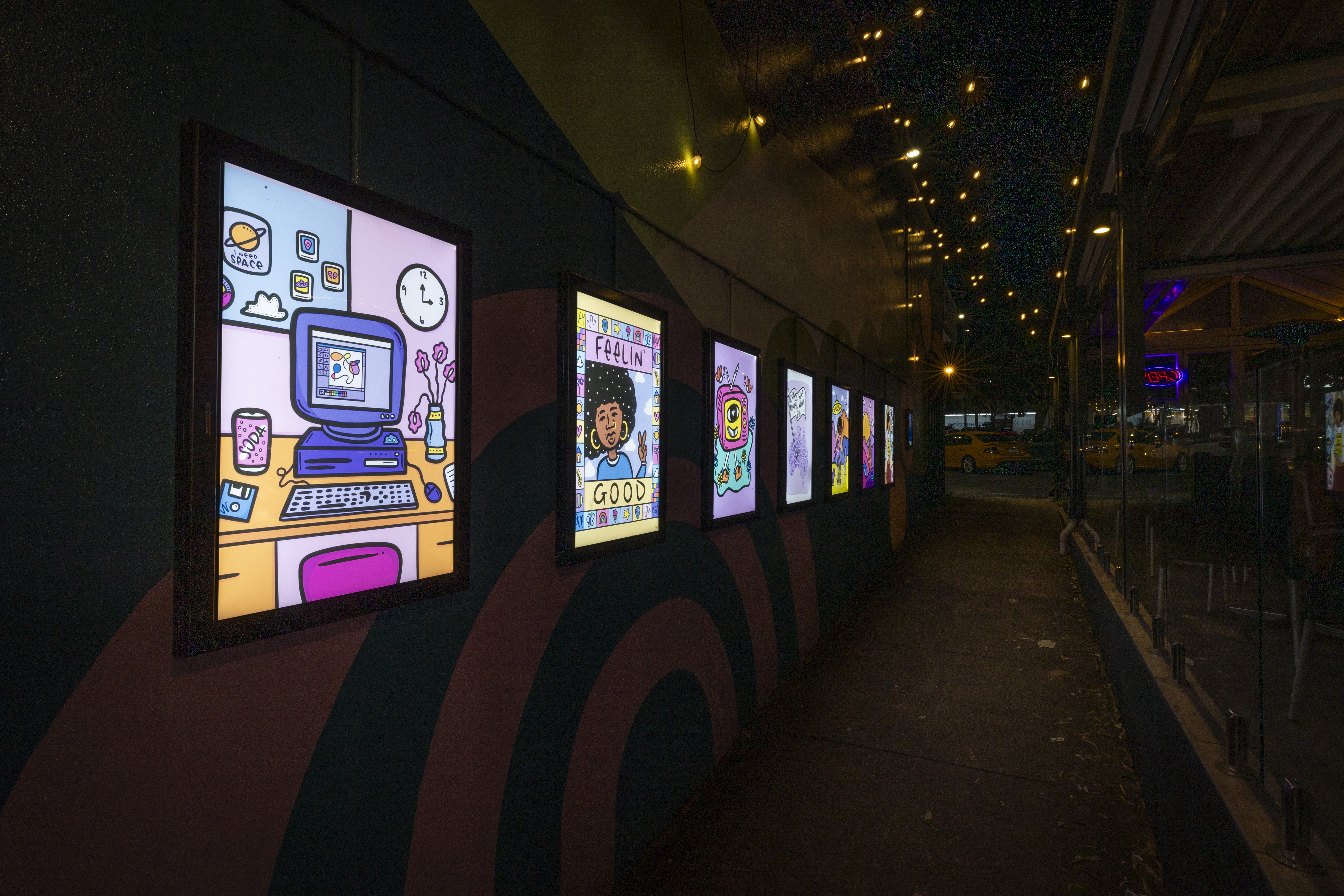 Night-time laneway art display featuring a series of illuminated digital artworks mounted along a wall, with decorative string lights overhead and reflections in nearby windows.