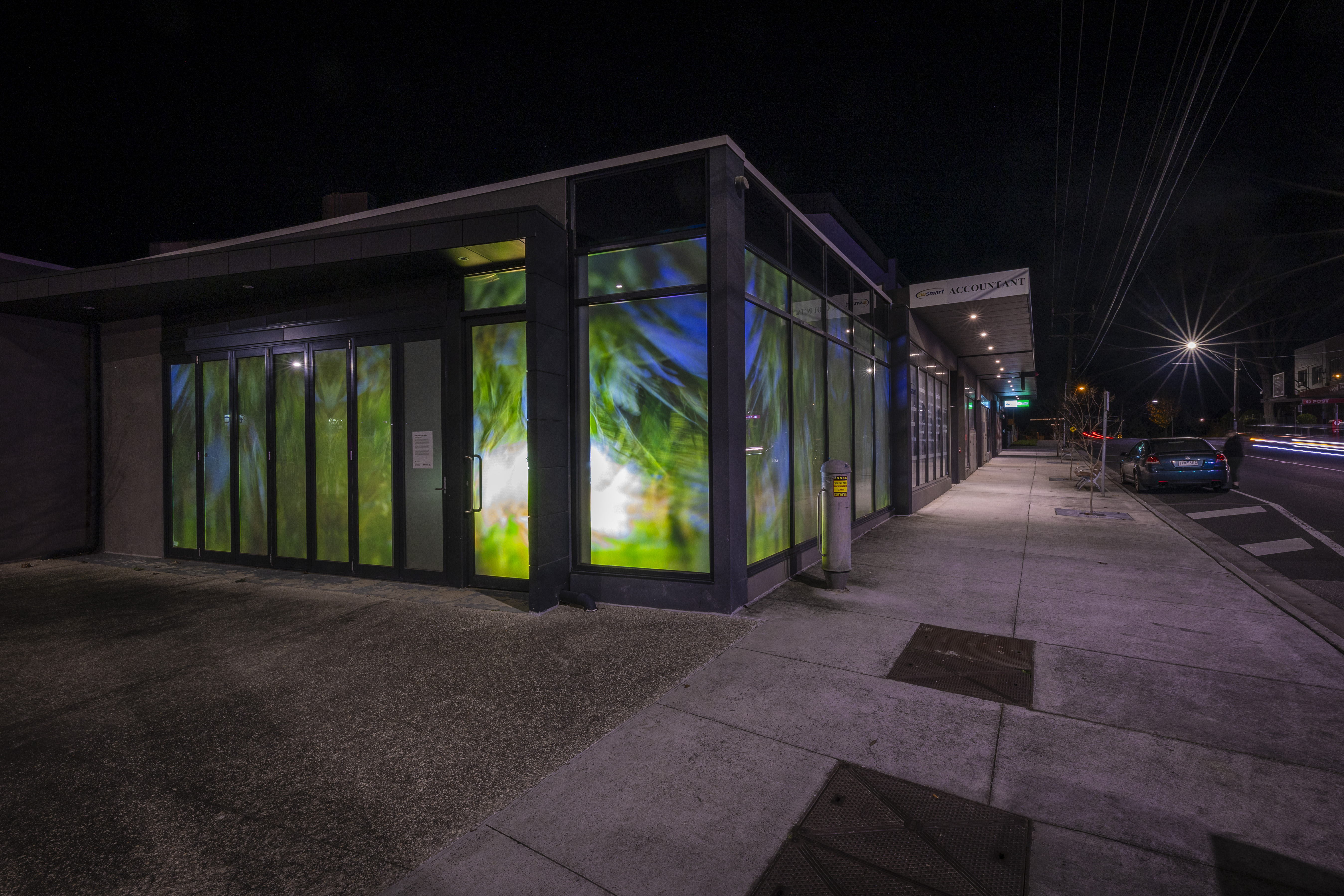 Contemporary corner building at night featuring illuminated window artwork in green, blue and white tones, casting colourful light onto a quiet streetscape.