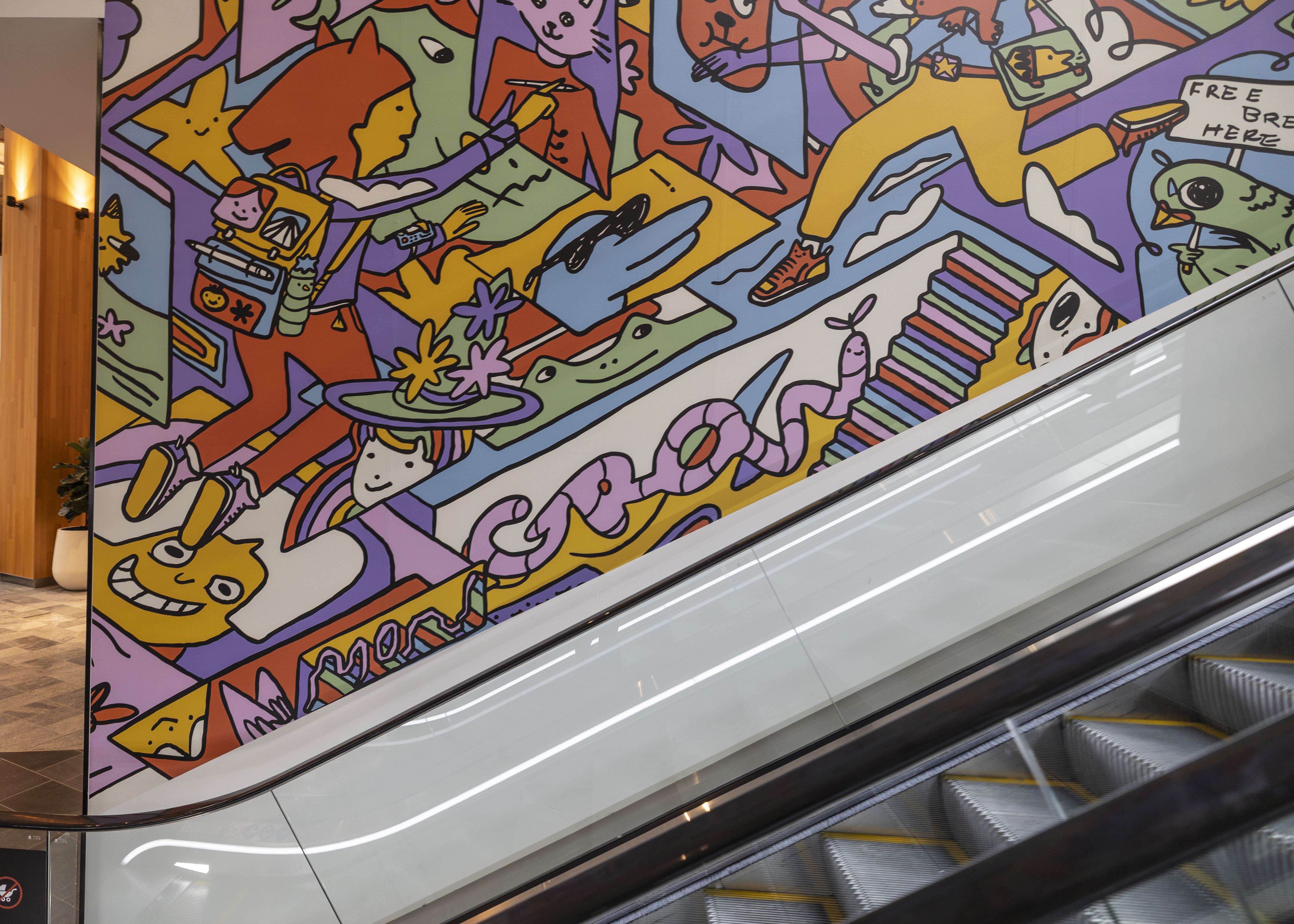 Large colourful mural beside an escalator, featuring playful cartoon-style characters, animals and abstract illustrations in bright pastel colours.