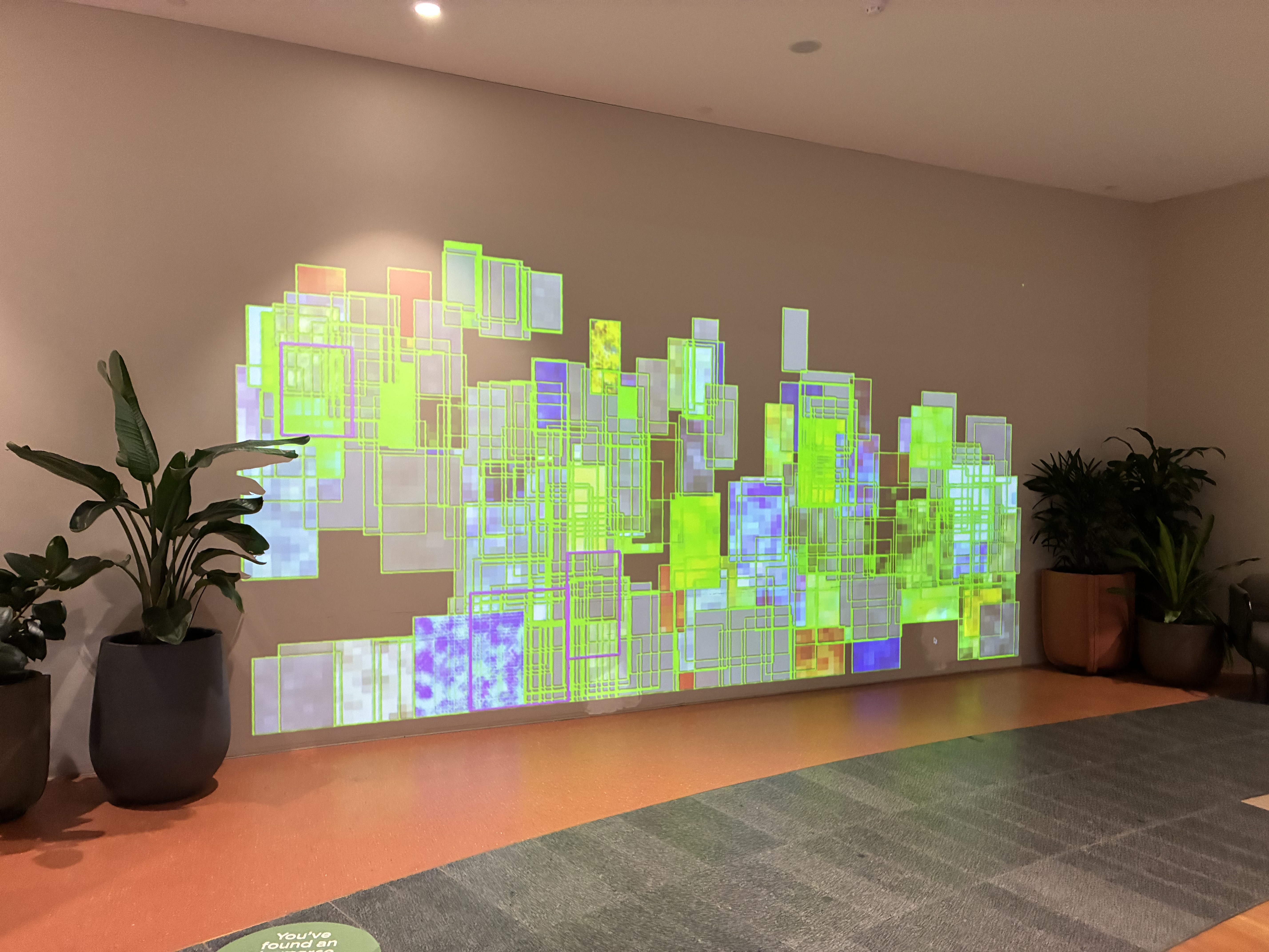 Indoor digital art installation projecting overlapping translucent rectangles in green, blue and purple tones across a large wall, framed by potted plants in a lobby space.