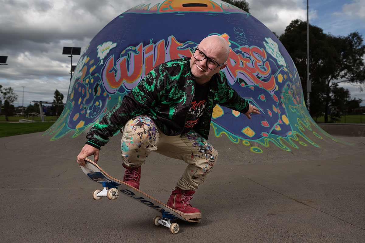 A man doing a trick on a skateboard on concrete in a skate park. He is in front of a painted mural on the skate bowl.