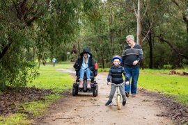 Photograph of a young male child on a bike, an adult male walking and an adult female in a wheelchair, travelling along a gravel path