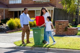 Two adults and young child on a residential street filling their red household rubbish bin