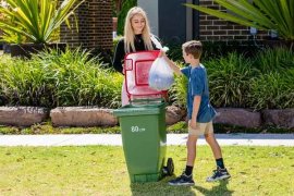 Young male and female on the street, placing rubbish bag in red household rubbish bin