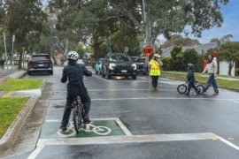 A school crossing supervisor helps an adult and child cross a local street. A cyclist waits in the foreground, and a car waits in the background.