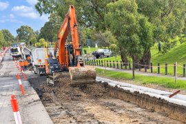 An orange excavator and construction workers dig a long trench beside a road, with traffic cones, trucks and a grassy, tree-lined park in the background.