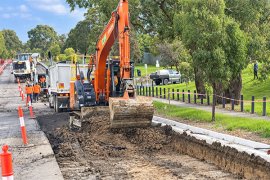 An orange excavator and construction workers dig a long trench beside a road, with traffic cones, trucks and a grassy, tree-lined park in the background.