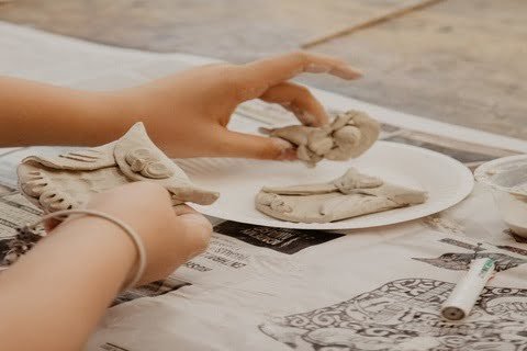 Picture of both of a child's hands moulding clay and using clay tools into the shape of an owl on a table covered in newspaper.