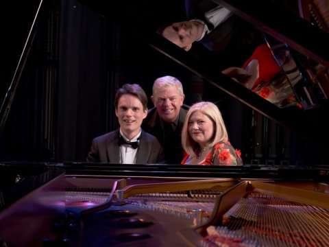 Picture of a black grand piano with the lid open and pianist man with dark hair, black suit bow tie at keyboard, older man in middle with greay hair and blonde woman with red top sitting on an angle and smiling toward the camera.