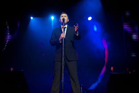 Man with short light brown hair standing at microphone stand, wearing dark suit with tie, left hand raised, singing, lights behind him on stage with lights in a blue and black hue.