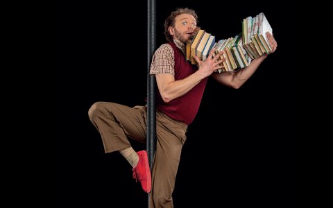 Man wearing red sneakers, brown slacks nad red vest with checkered red shirt, looking wide eyed and shocked, with light brown hair and beard with his leg slung around a black dance pole holding approx 15 books in his right arm and balancing it under his chin on a black background.