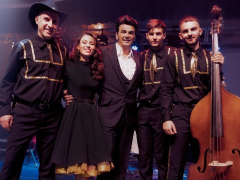 Band of four men and one girl with long red brown flowing hair standing closely togethe with arms around eachothers waists wearing black clothing gold trim. Man on left is wearing a black cowboy hat and man on right is holding a cello on the floor and the neck in his left hand