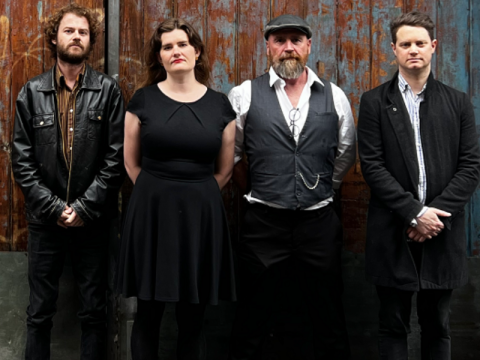 the four band members, three men and one woman, standing infront of an old worn timber gate of red brown and grey colour, looking straight at the camera wearing dark clothing. Gaille the band lead has a black riders cap on and white shirt with black vest.