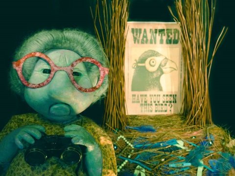 Puppet of an older person with grey hair and big red round glasses, with lighting in shade of blue and green, holding binoculars and standing next to a table with straw and a black and white sign with a poster saying wanted with a picture of a birds head on it.