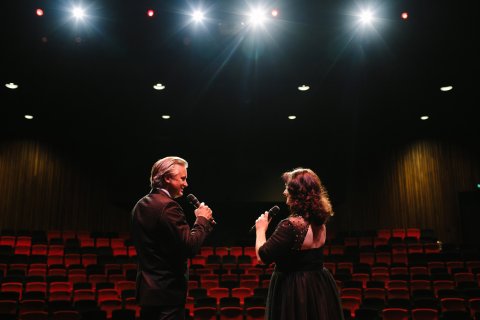 Man with light coloured hair holding microphone to mouth with woman with long  dark red hair looking at each othe with back turned to camera, in a theatre with bright lights above them and empty rows of red theatre seats in front of them.