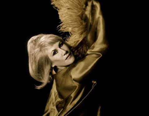 Lady with shoulder length light blonde hair with arms raised above her head, standing on an angle to the right looking into the camera, picutre from waste up wearing a gold satin gown holding a feather boa in the air, background is black.