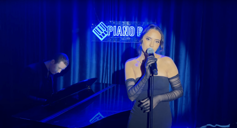 You lady with strapless tight black dress, sheer arm length gloves and dark hair pulled back, holding the microphone on a stand in her right hand and left hand on the stand, singing softly with head tilted with blue spotlight on her and man on piano playing keyboards to her left with a blue luminous sign again the dark curtain in the background with Pioan Bar highlighted on it.