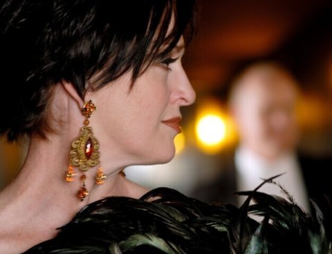 Lady with short dark brown hair and gold and red chandelier earrings, standing side on looking to the right, head and shoulders only, holding a black feather boa to her chest