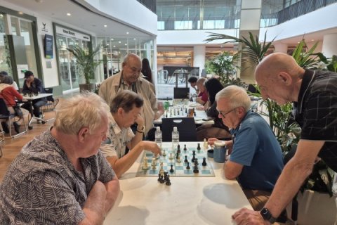 Group of people gathered around a table in a bright indoor atrium, focused on a chess game. Several people sit while two others stand and observe. The space includes glass walls, plants, café seating, and other people in the background.