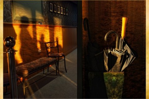 Split scene showing a warmly lit interior: on the left, a wooden bench and small chair against a wall are illuminated by low sunlight casting long shadows; on the right, several umbrellas with curved handles rest in a stand beside a textured wall.