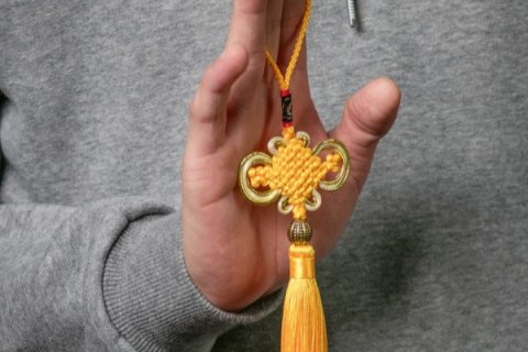 Close-up of a hand holding a decorative yellow knot ornament with a tassel, suspended by a cord. The person is wearing a grey top, and the detailed woven pattern of the ornament is clearly visible.