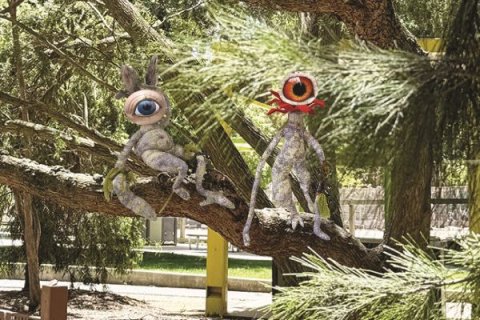 Two small, surreal creature sculptures with oversized single eyes sit on the branch of a tree in an outdoor setting. One figure reclines against the branch while the other stands upright; both have textured, handmade surfaces. Pine needles and branches frame the scene, with buildings and a walkway visible in the background.