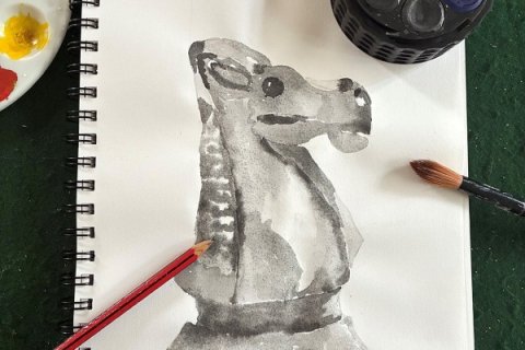 Overhead view of an open sketchbook on a green work surface showing a monochrome painting of a horse or knight chess piece rendered in grey wash. A red pencil rests diagonally across the page, while a paintbrush lies to the right. At the top edge, a small paint palette with yellow paint and a dark container are partially visible, reinforcing an active art-making setup.