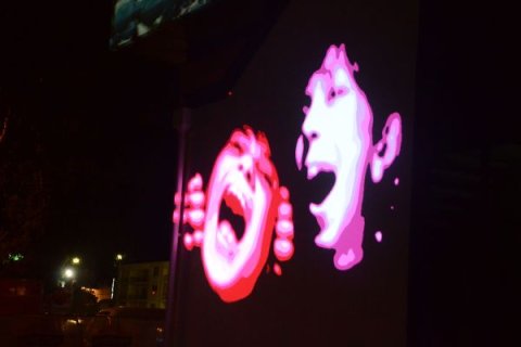 Night-time scene showing a building exterior with two large, brightly coloured projected faces on the wall. The faces appear stylised and partially overlapping in red, pink, and white tones, standing out against the dark surroundings.