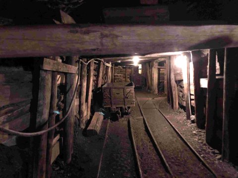 Interior view of an underground coal mine with timber supports, narrow rail tracks, a mining cart, and dim overhead lighting extending into the tunnel.