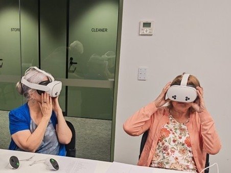 Two people seated at a table indoors wearing virtual reality headsets during a technology-based learning or recreation session.