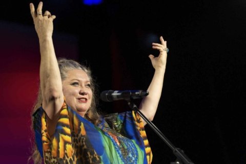 Lady Shaula standing at a microphone on stage with both arms raised, wearing a brightly coloured patterned outfit against a dark background illuminated by stage lighting.